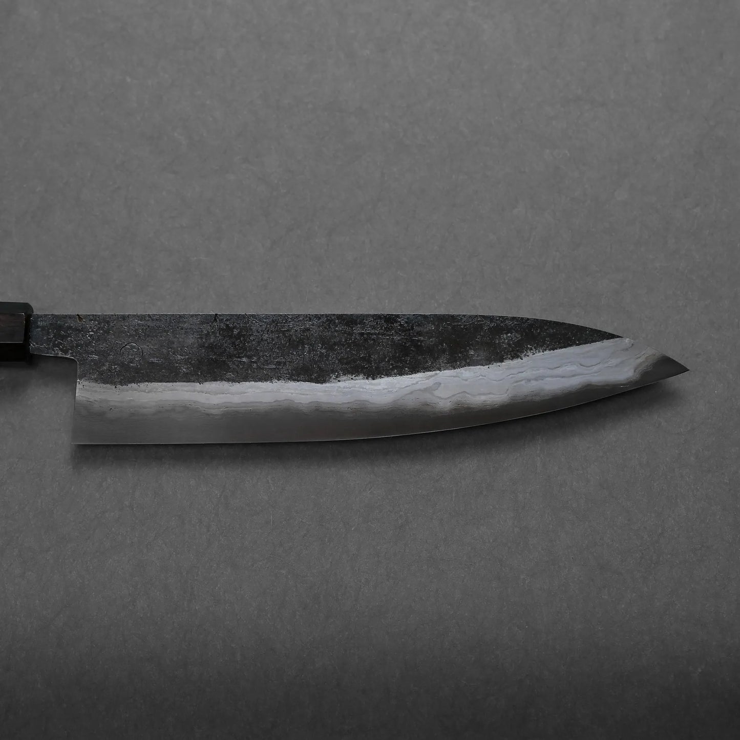 A close-up view of a chef's knife with a dark, textured Damascus blade and a visible wavy pattern along the edge, set against a plain gray background.