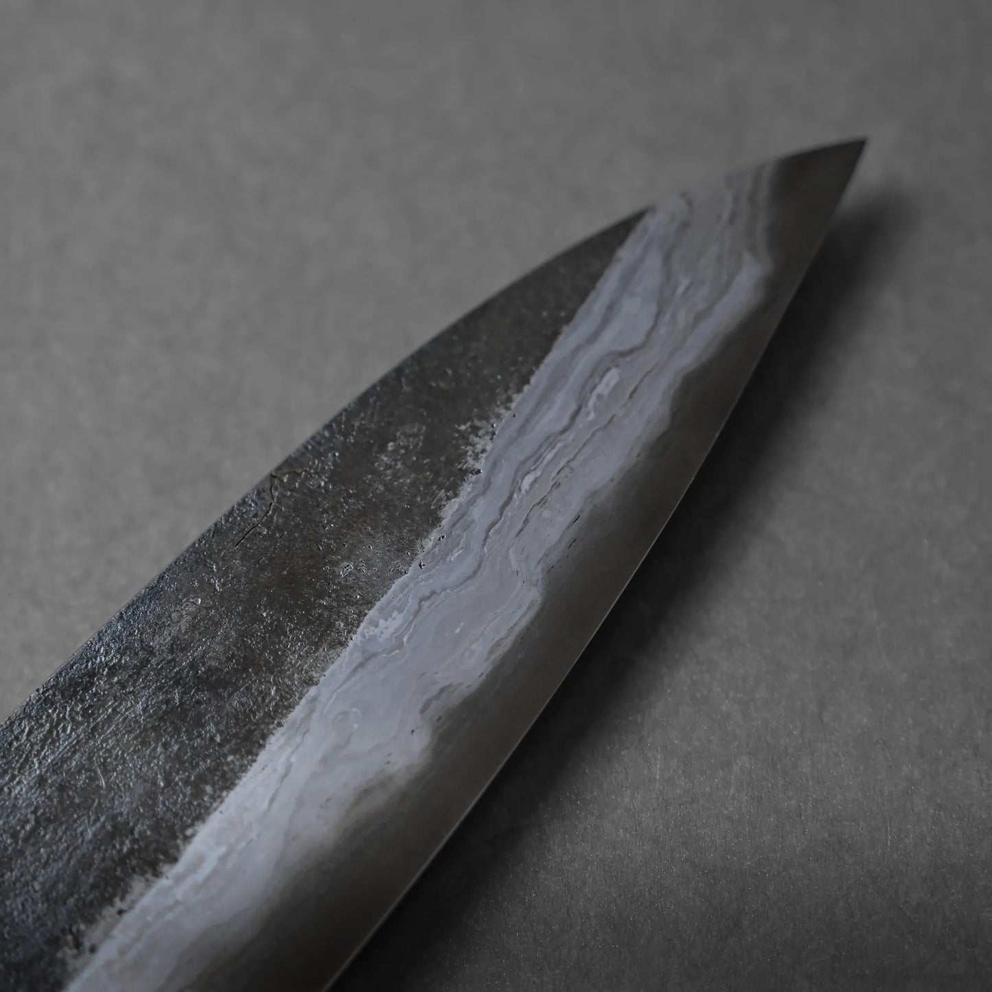Close-up of a dark, textured Damascus knife blade with a visible wavy pattern along the edge, set against a plain gray background.