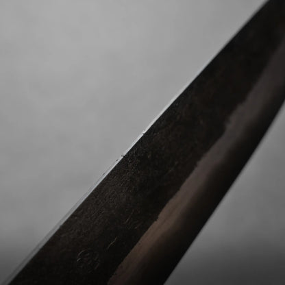 A close-up image of the edge of a dark metal knife blade, showing a small chip or imperfection along the worn metal. The background is out of focus and gray.