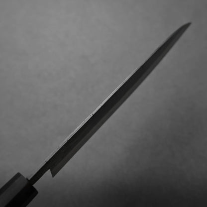 A close-up, angled view of a kitchen knife blade against a plain gray background. The focus is on the thin edge of the blade, highlighting its metallic texture and emphasizing its precise craftsmanship.