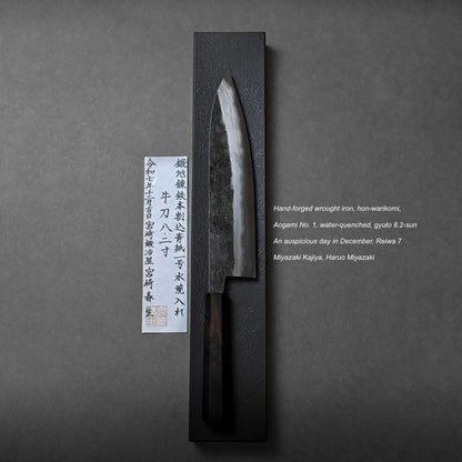 A hand-forged Japanese chef’s knife with a dark blade and handle is displayed vertically on a black stand. A certificate with Japanese text and a brief English description accompanies the artisanal knife to the left.