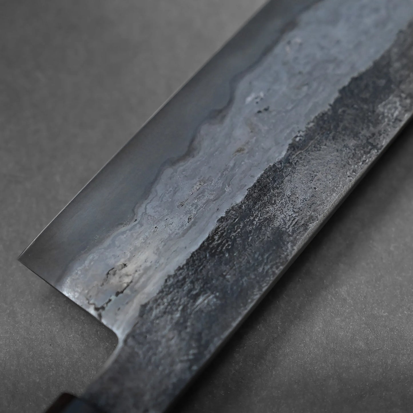 Close-up of a Miyazaki rentetsu gyuto blade shows intricate patterns and textures on the metal, with a textured area near the edge and a smoother polished section above. The background is plain and dark.