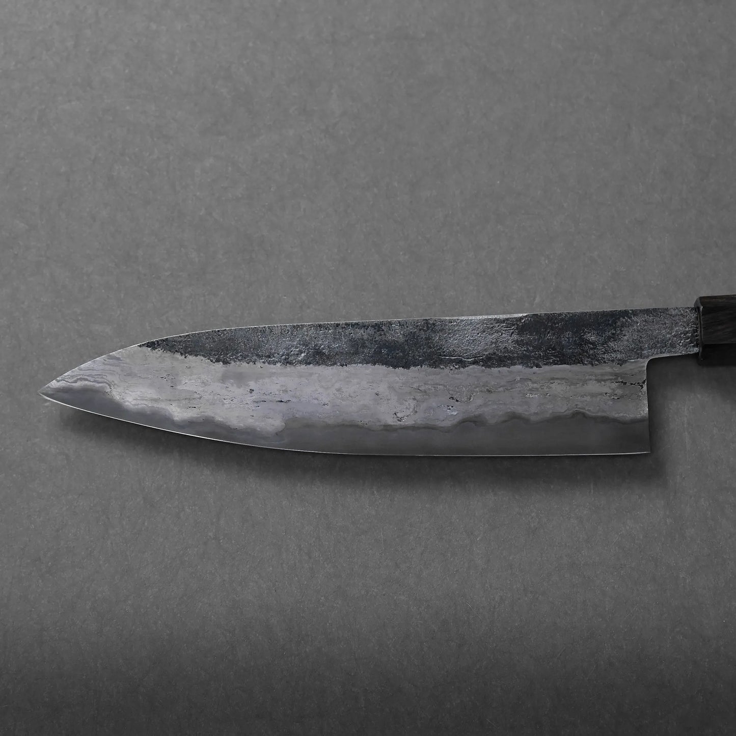 A close-up of a Miyazaki rentetsu gyuto Japanese-style kitchen knife with a dark, weathered blade and textured patterns, photographed on a plain gray surface.