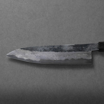 A close-up of a Miyazaki rentetsu gyuto Japanese-style kitchen knife with a dark, weathered blade and textured patterns, photographed on a plain gray surface.