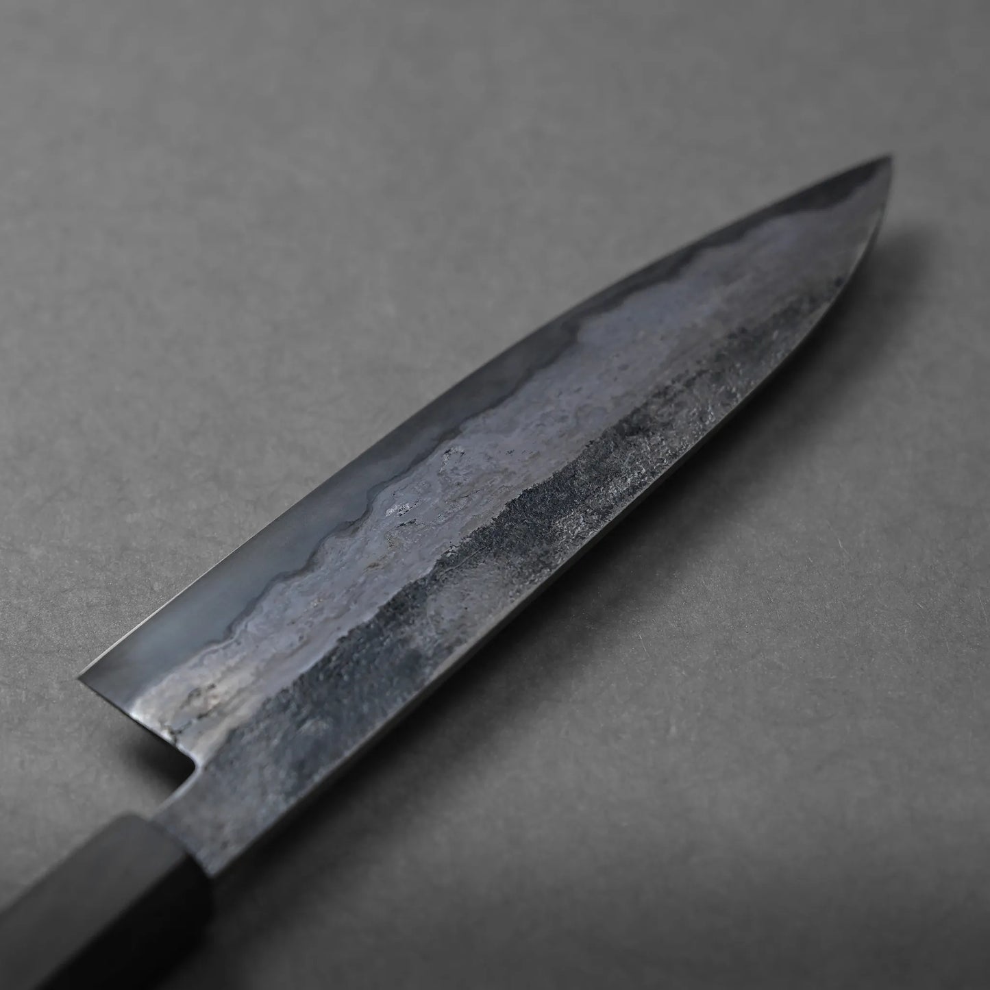 A close-up view of a Miyazaki rentetsu gyuto Japanese-style chefs knife with a dark, textured steel blade and a black handle, placed on a plain gray surface.