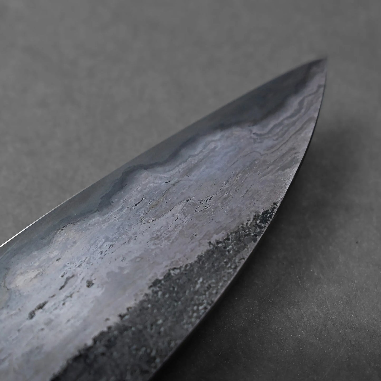 A close-up view of the pointed tip of a Miyazaki rentetsu gyuto blade, showing detailed patterns and textures in the steel against a plain gray background.