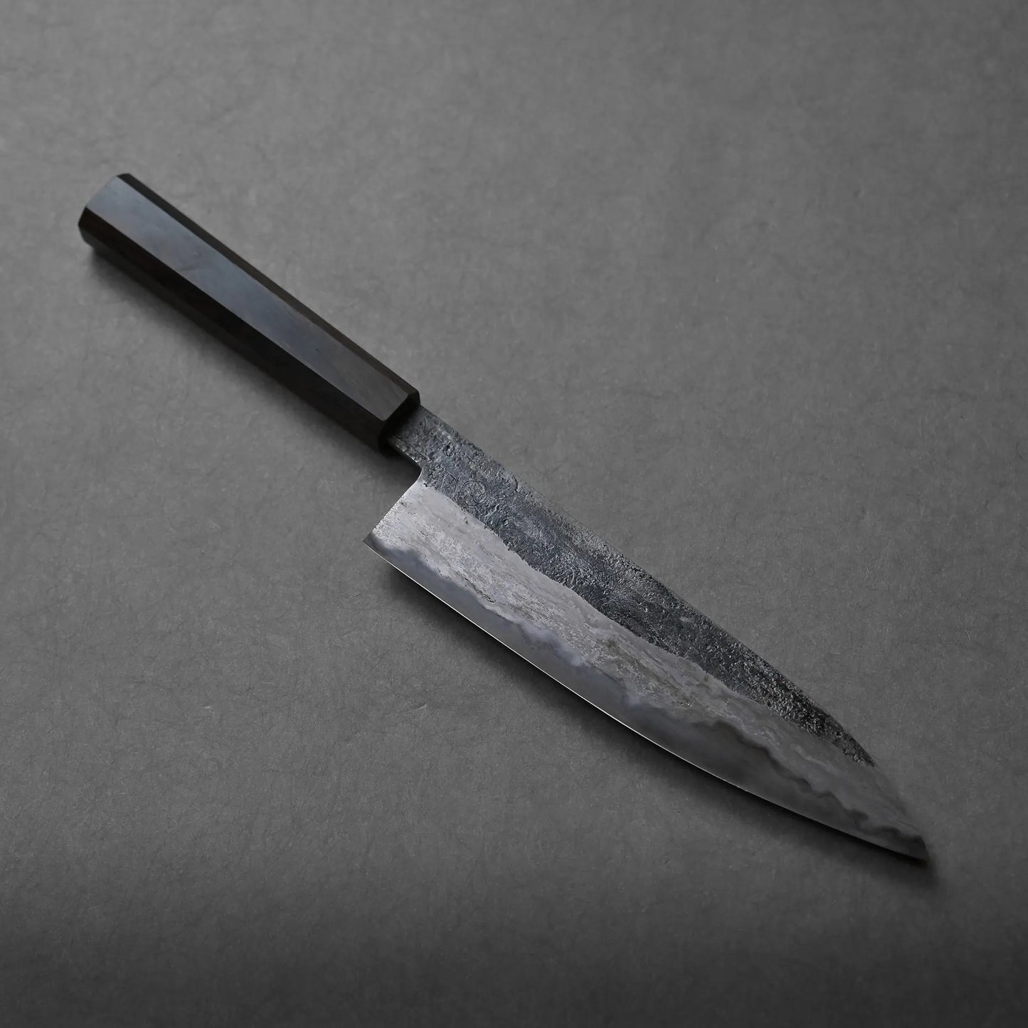 A handcrafted Miyazaki rentetsu gyuto kitchen knife with a dark wooden handle and a textured, partially polished blade rests on a gray surface.