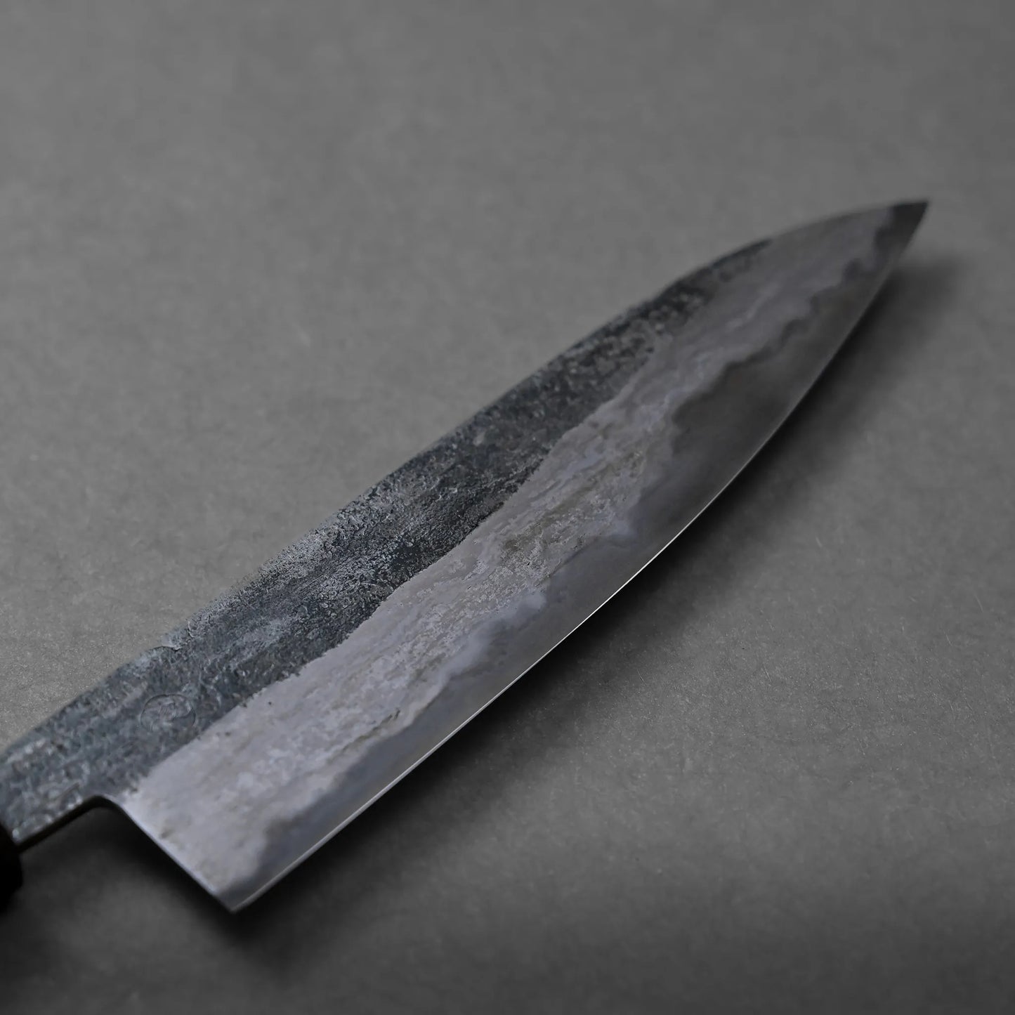 A close-up view of a large, dark Miyazaki rentetsu gyuto kitchen knife with a textured surface, placed on a plain gray background.