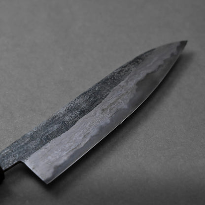 A close-up view of a large, dark Miyazaki rentetsu gyuto kitchen knife with a textured surface, placed on a plain gray background.
