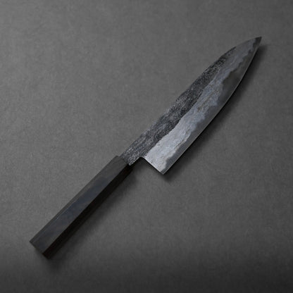 A Miyazaki kurouchi rentetsu shirogami#1 gyuto 210mm with a dark, textured blade and a rectangular handle rests on a grey surface.