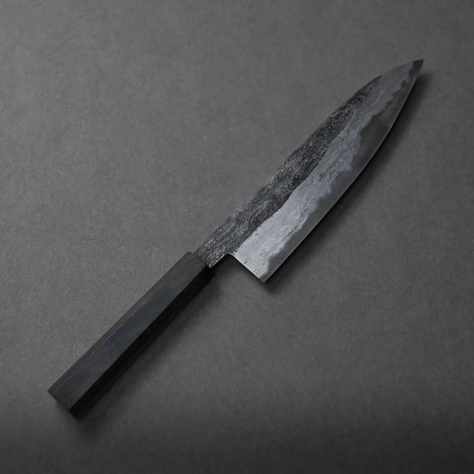 A Miyazaki kurouchi rentetsu shirogami#1 gyuto 210mm with a dark, textured blade and a rectangular handle rests on a grey surface.
