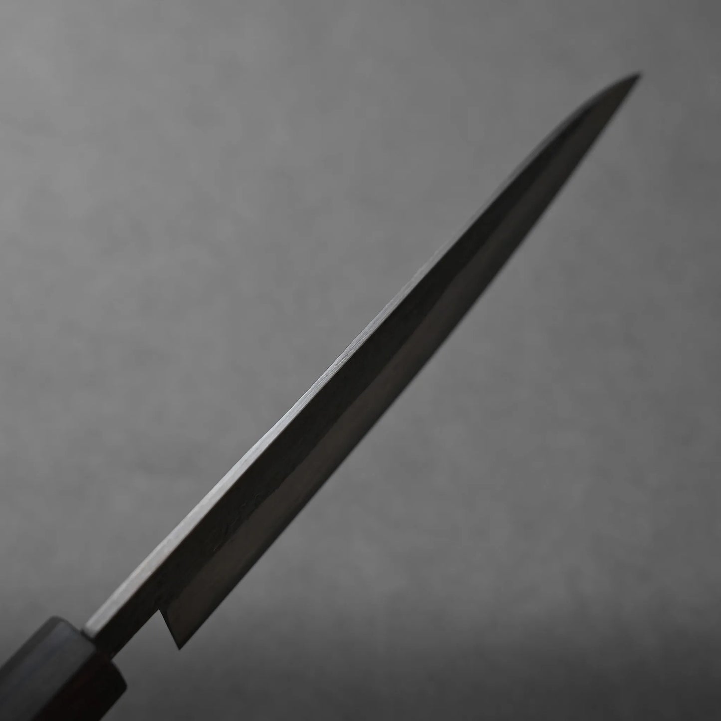 A close-up of a Miyazaki rentetsu gyuto kitchen knife with a dark handle, set against a plain gray background. The blade is angled diagonally and occupies most of the image.