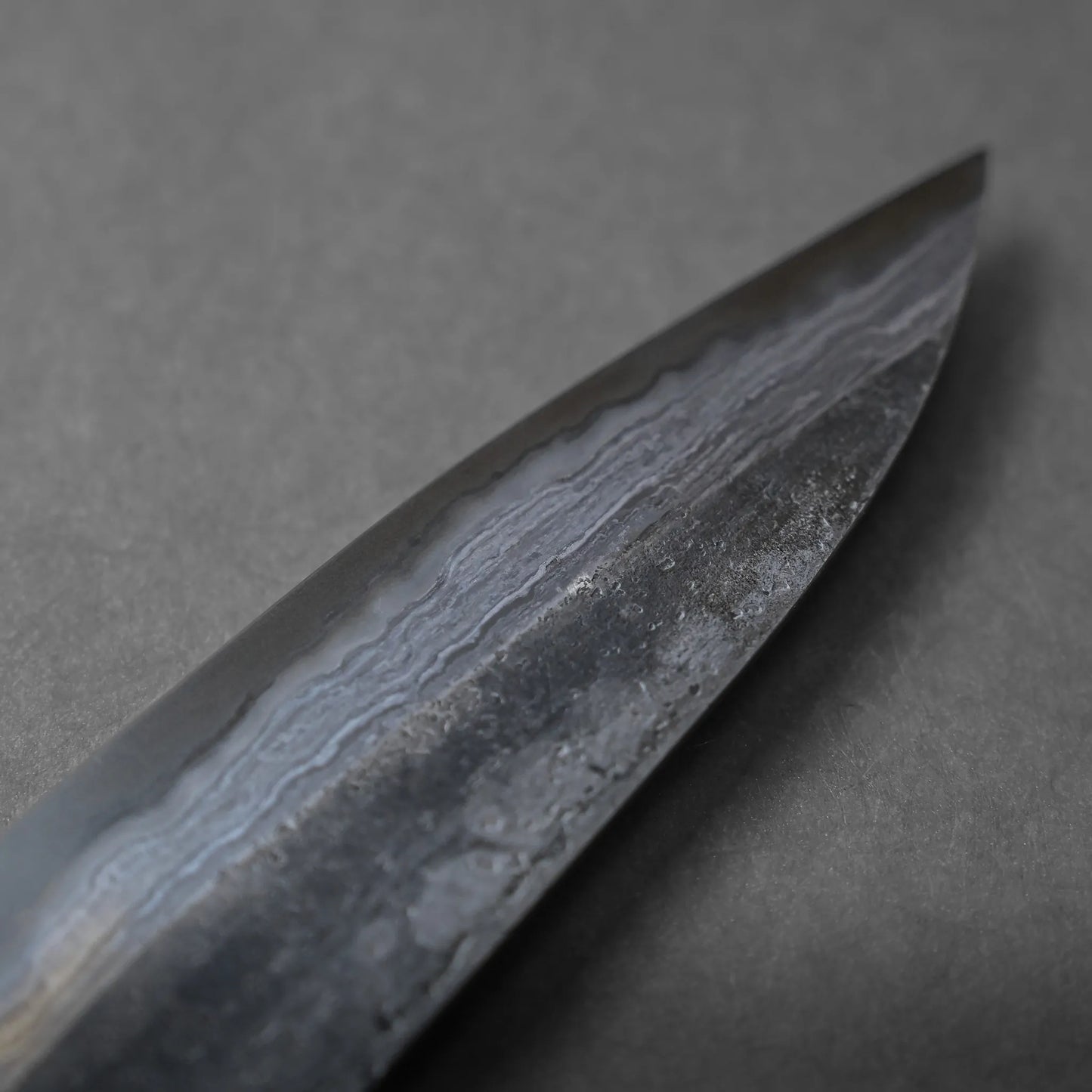 Close-up of the tip of a Damascus steel knife blade, displaying intricate wavy patterns and unique texture on the metal surface, photographed against a plain dark background.