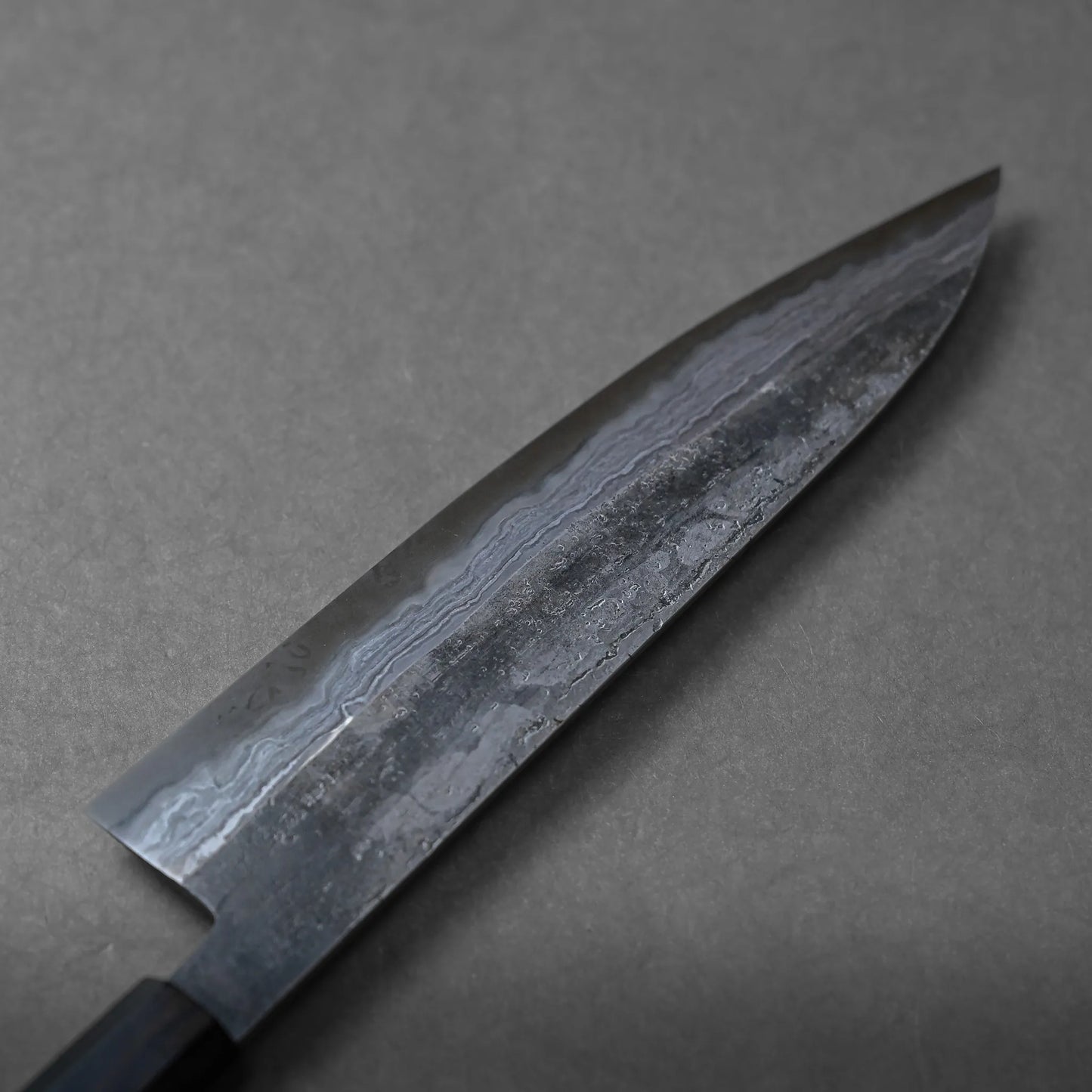 A close-up of a Japanese chef’s knife with a black handle, placed on a gray surface. The blade features striking Damascus-style wave patterns, highlighting the traditional craftsmanship.