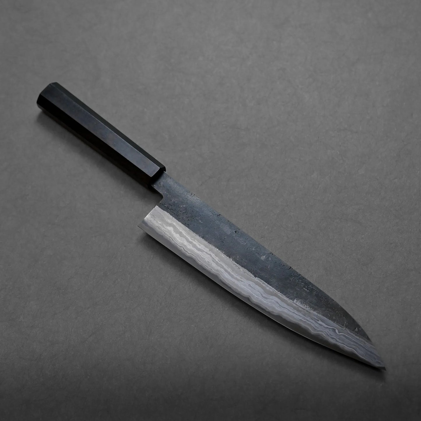 A Japanese-style kitchen knife with a dark wooden handle and a patterned, slightly reflective blade is displayed on a gray surface. Intricate wavy patterns near the edge highlight its artisanal craftsmanship.