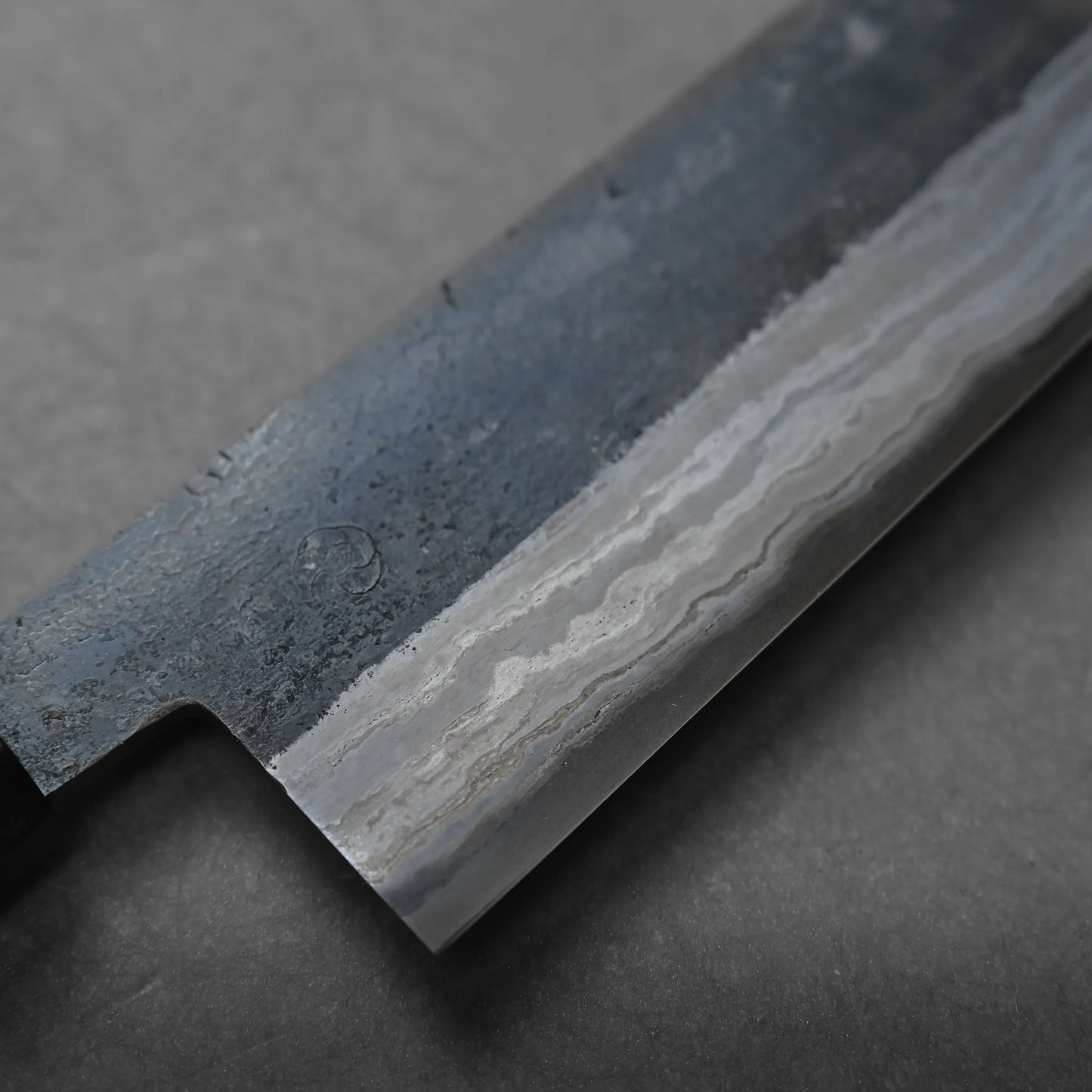 A close-up view of a Japanese chefs knife blade reveals a distinctive wavy pattern characteristic of Damascus steel against a gray background, highlighting its craftsmanship.