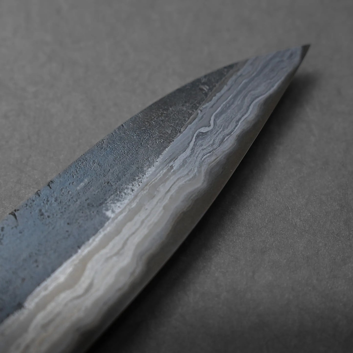 Close-up of the tip of a Damascus steel knife blade, highlighting unique wavy patterns and intricate textures on the metal, set against a neutral gray background.