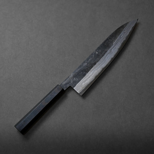 A Miyazaki kurouchi rentetsu shirogami#1 gyuto 245mm with a dark wooden handle and a patterned, slightly reflective blade rests on a plain, dark gray surface, showcasing its artisanal craftsmanship.