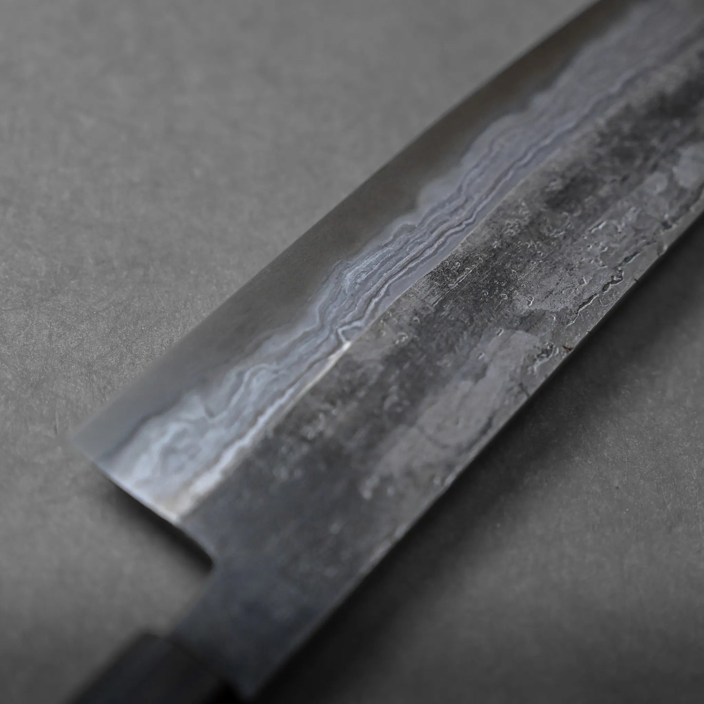 Close-up of a Japanese chefs knife blade, featuring intricate Damascus patterns and unique texture on the metal surface, set against a gray background.