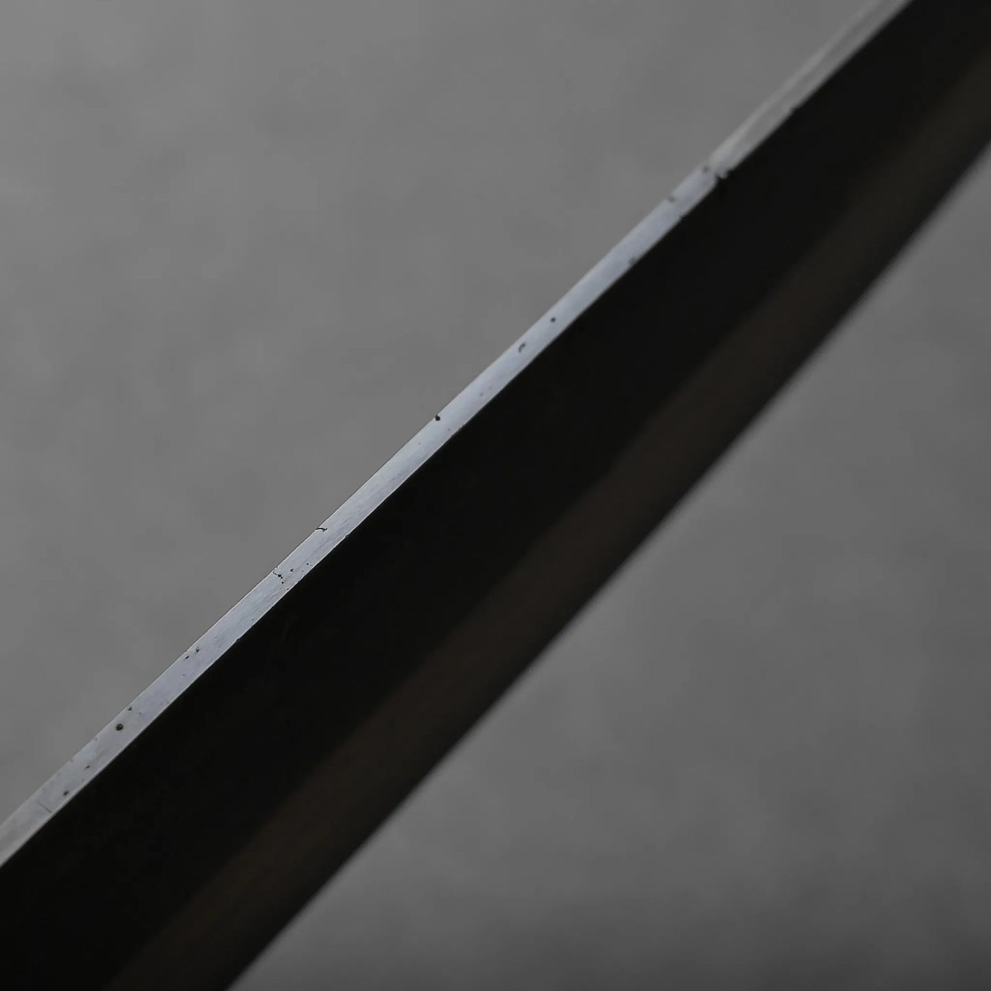A close-up view of the metallic blade's edge against a plain, neutral gray background. The metal surface reflects light, highlighting fine details along the blade.