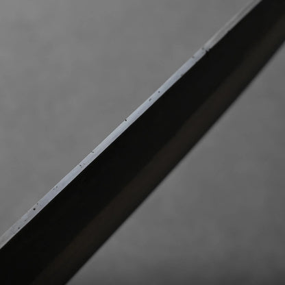 A close-up view of the metallic blade's edge against a plain, neutral gray background. The metal surface reflects light, highlighting fine details along the blade.
