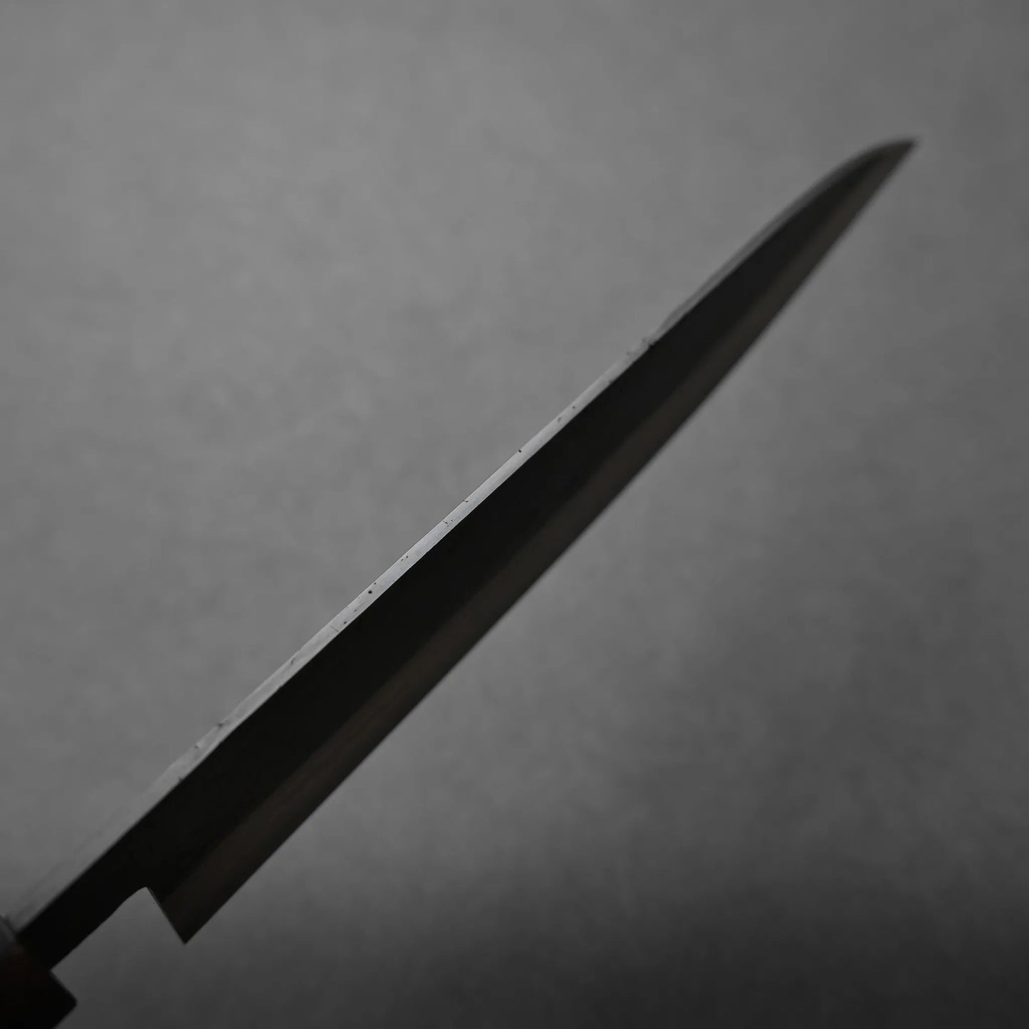 A close-up view of a kitchen knife blade set against a plain, gray background. The blade is angled diagonally across the image, showcasing its thin, pointed edge.