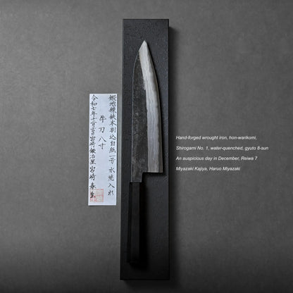 A hand-forged Japanese kitchen knife displayed vertically on a black rectangular stand, accompanied by a paper with Japanese calligraphy and English text detailing the artisan, materials, and creation date.