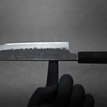 A hand wearing a black glove balances a large Japanese chef's knife on the tip of an upright index finger against a plain gray background. It shows the balance point.