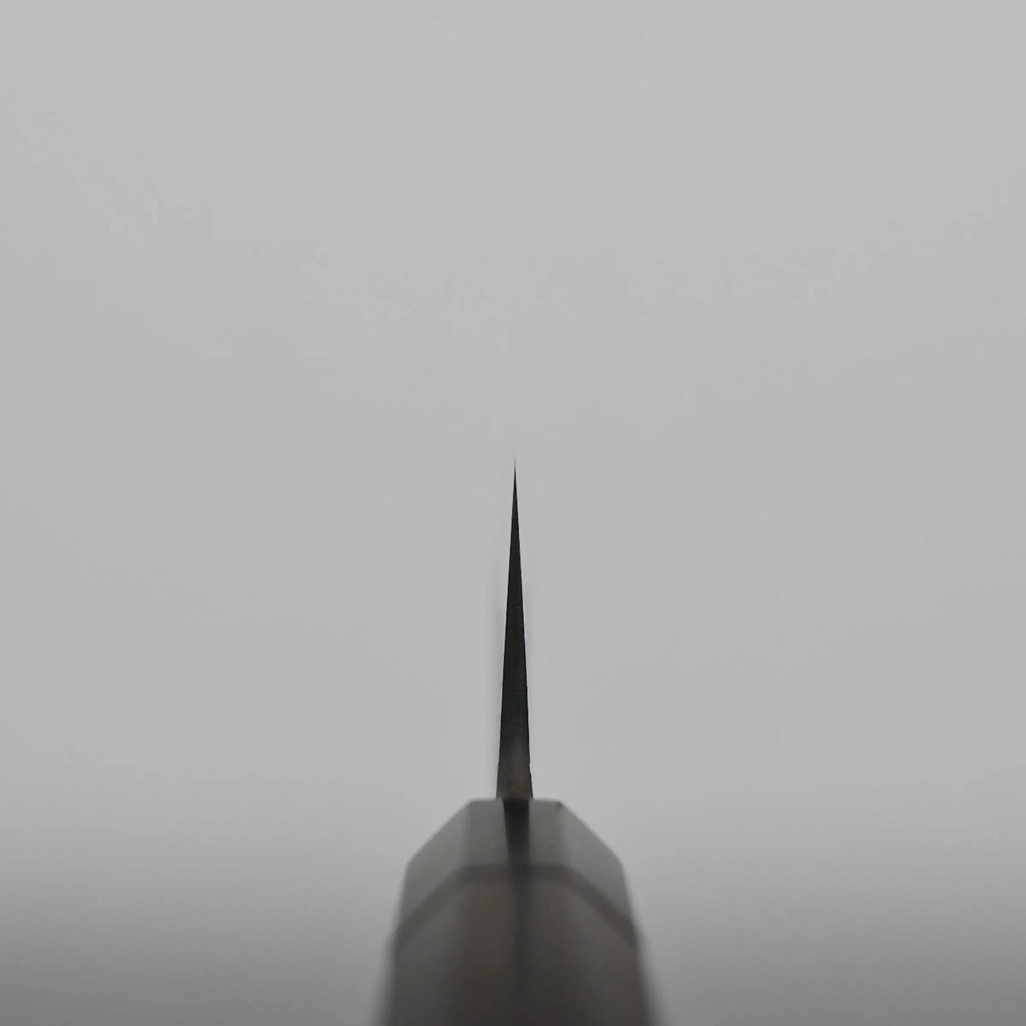 A close-up image of the tip of a Japanese chef's knife blade photographed from a straight-on angle against a plain, light gray background. The blade tapers to a fine point and is centered in the frame.