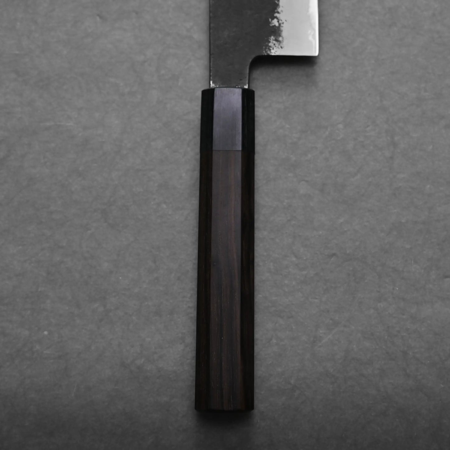 Close-up of the handle and part of a gyuto knife, shown against a gray textured background. The dark brown wooden handle has visible grain, and the blade of this Japanese chef's knife has a slightly reflective, unfinished edge.