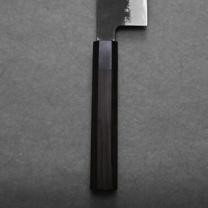 Close-up of the handle and part of a gyuto knife, shown against a gray textured background. The dark brown wooden handle has visible grain, and the blade of this Japanese chef's knife has a slightly reflective, unfinished edge.