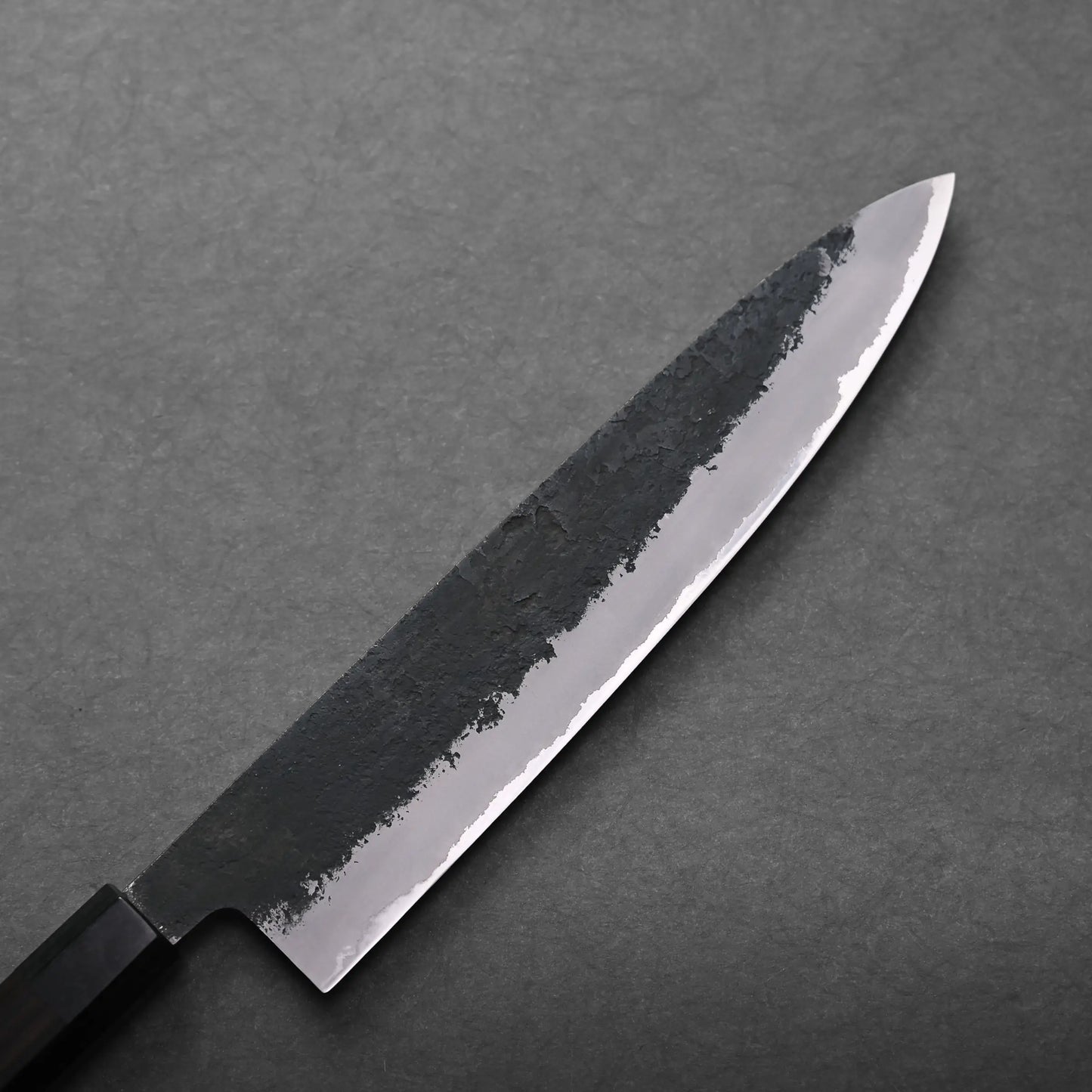 A close-up of a Japanese chef's knife with a dark, textured blade and a straight-edged handle, placed on a gray surface.