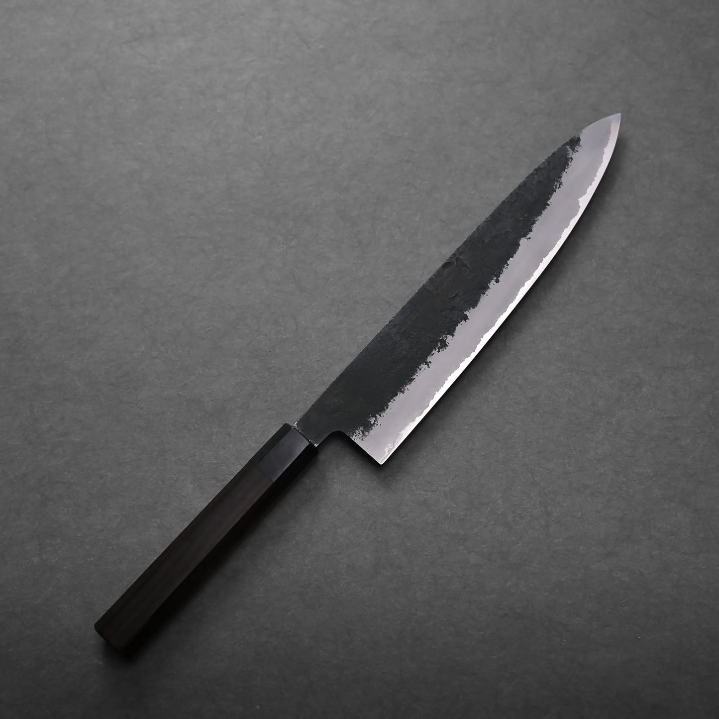 A Mutsumi Hinoura kurouchi nashiji AS gyuto 240mm with a dark wooden handle and a partially polished, partially matte blade rests on a dark gray surface.