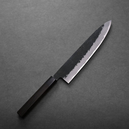A Mutsumi Hinoura kurouchi nashiji AS gyuto 240mm with a dark wooden handle and a partially polished, partially matte blade rests on a dark gray surface.