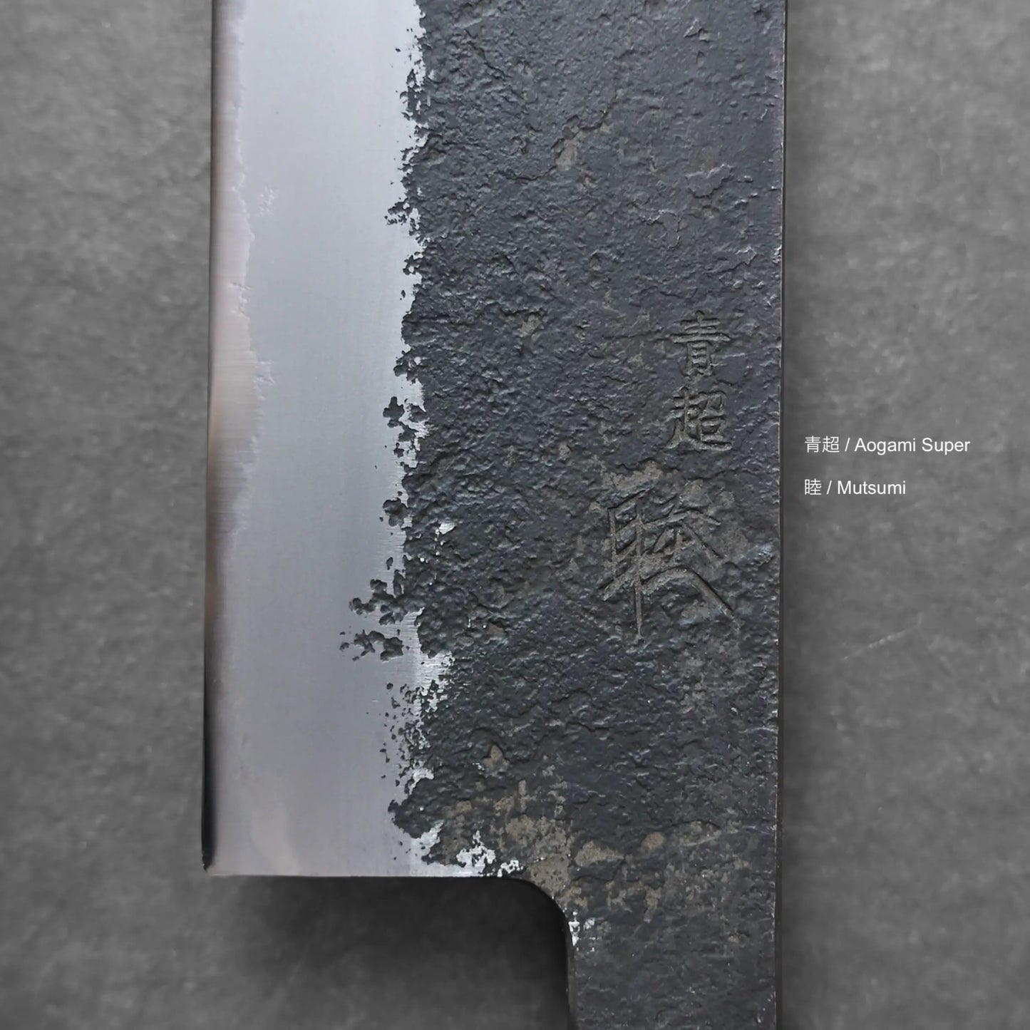 Close-up of a Japanese chef's knife blade with a textured, dark rough area near the edge and polished steel above. Japanese characters and the words Aogami Super and Mutsumi are engraved on this gyuto knife.