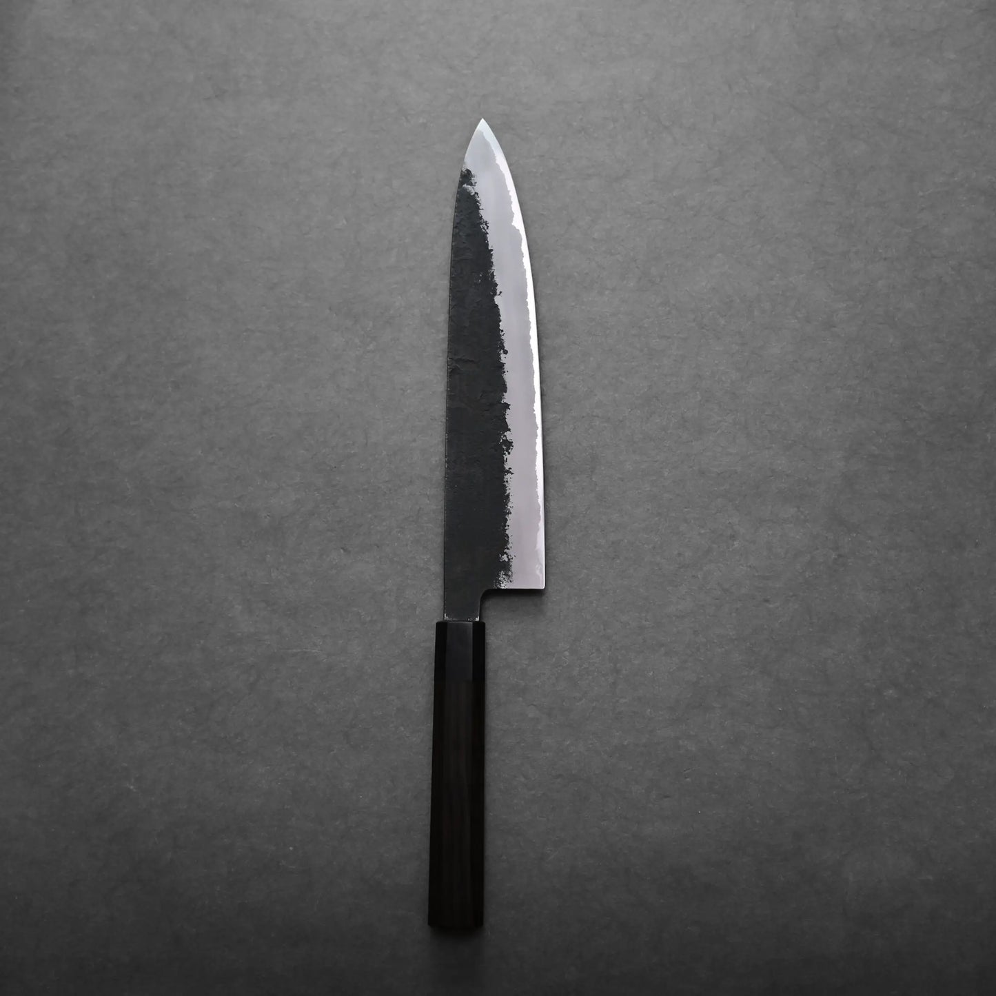 A single gyuto knife with a dark wooden handle and a polished, slightly textured blade rests on a gray surface.