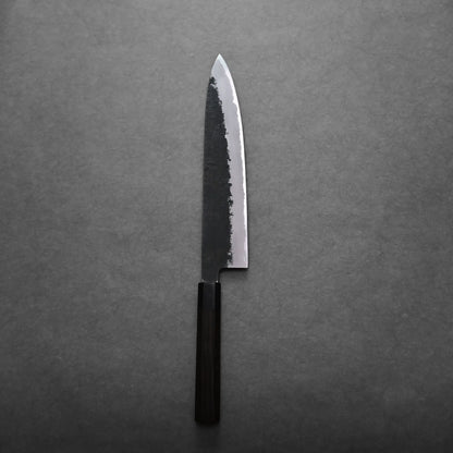 A single gyuto knife with a dark wooden handle and a polished, slightly textured blade rests on a gray surface.