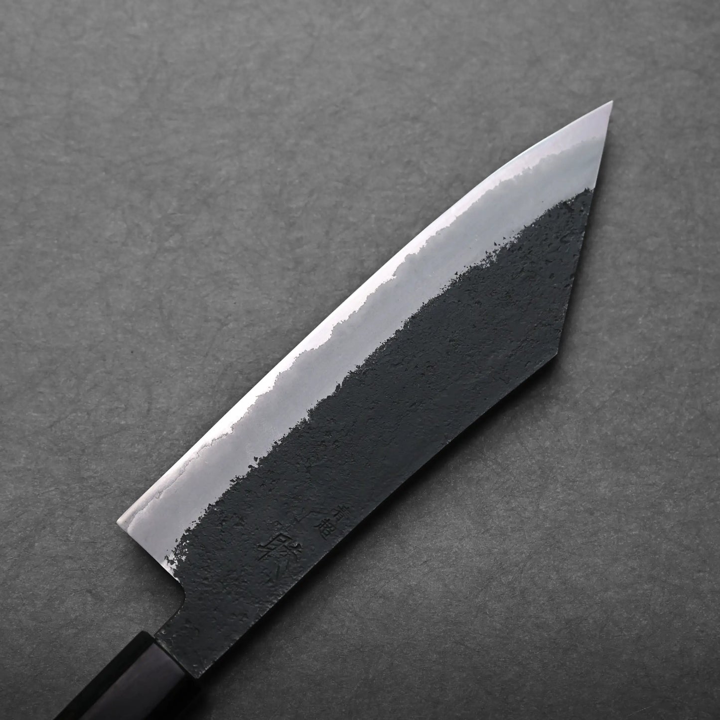 A close-up view of a Japanese-style hakata knife with a black, textured blade and sharp, pointed tip, placed on a gray surface.