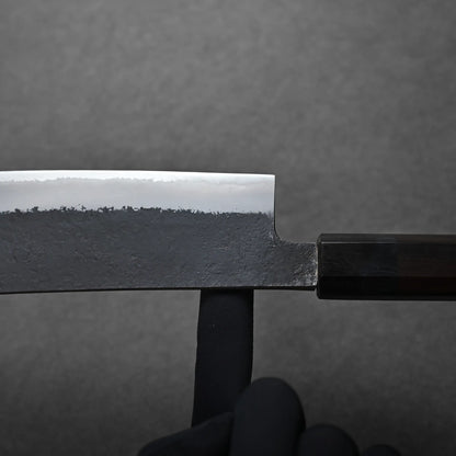 A hand in a black glove balances a Hakata knife on one finger against a plain dark gray background. The knife features a shiny, polished blade with a contrasting textured, dark lower section and a black handle. It shows the balance point.