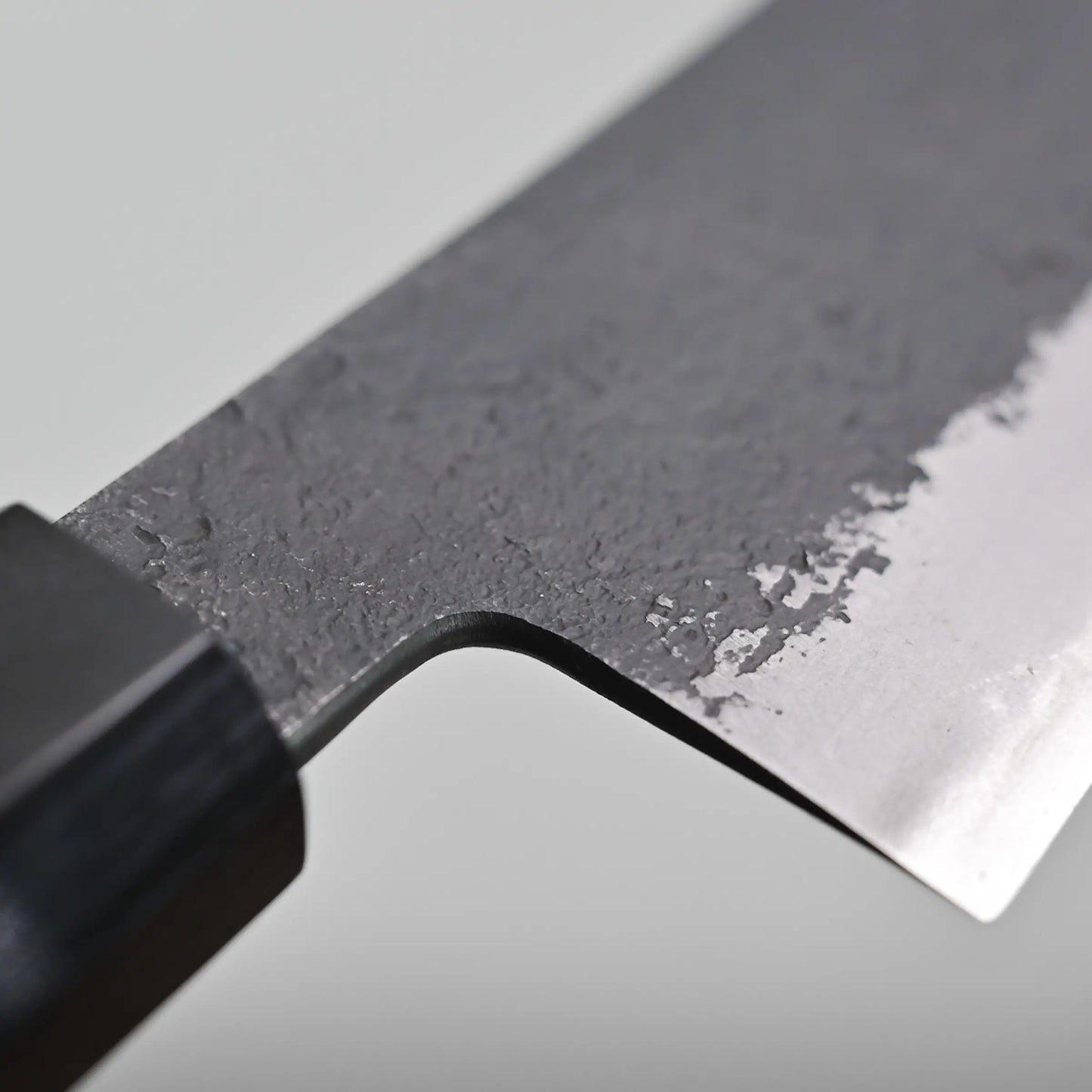 Close-up of a hakata knife blade revealing a textured, dark upper portion and a smooth, sharp lower edge, with part of the handle visible on the left.