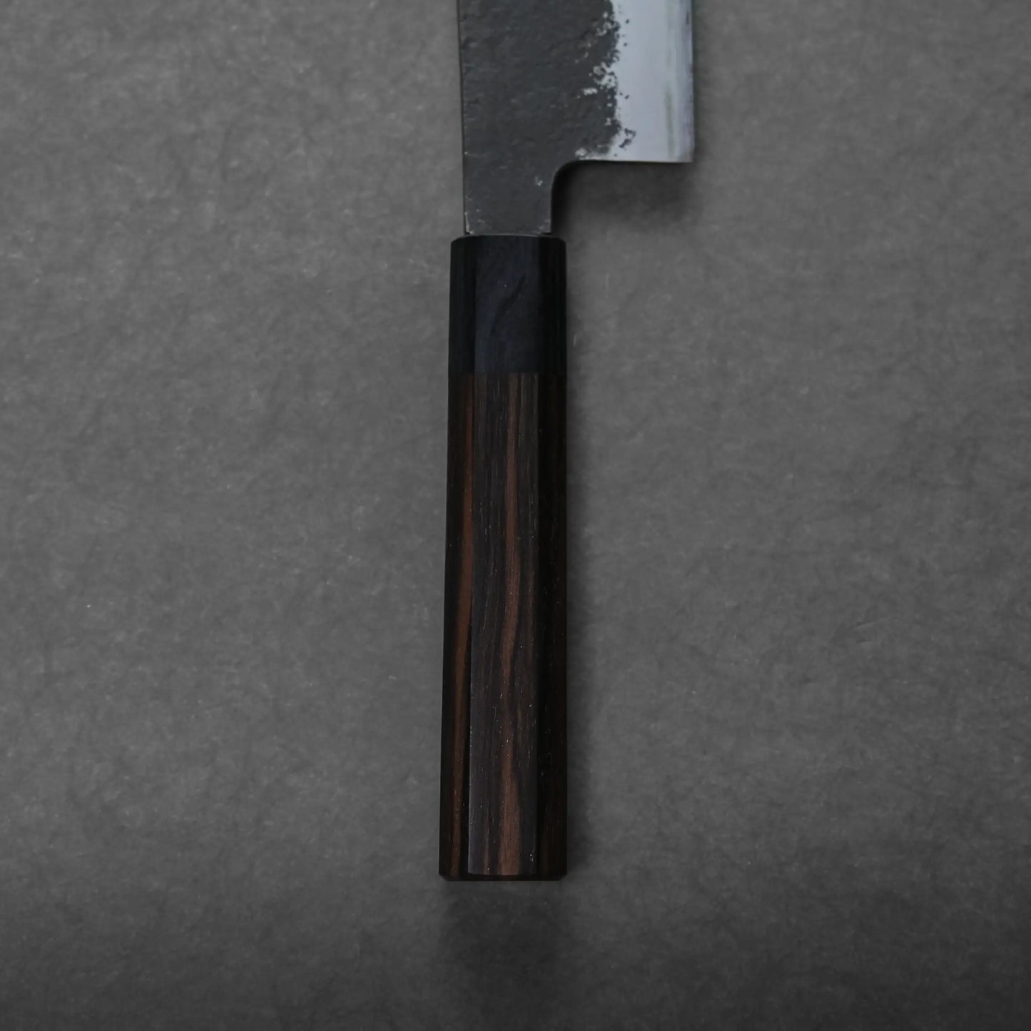 A close-up of a hakata knife with a dark wooden handle and a partially visible blade on a gray, textured surface.