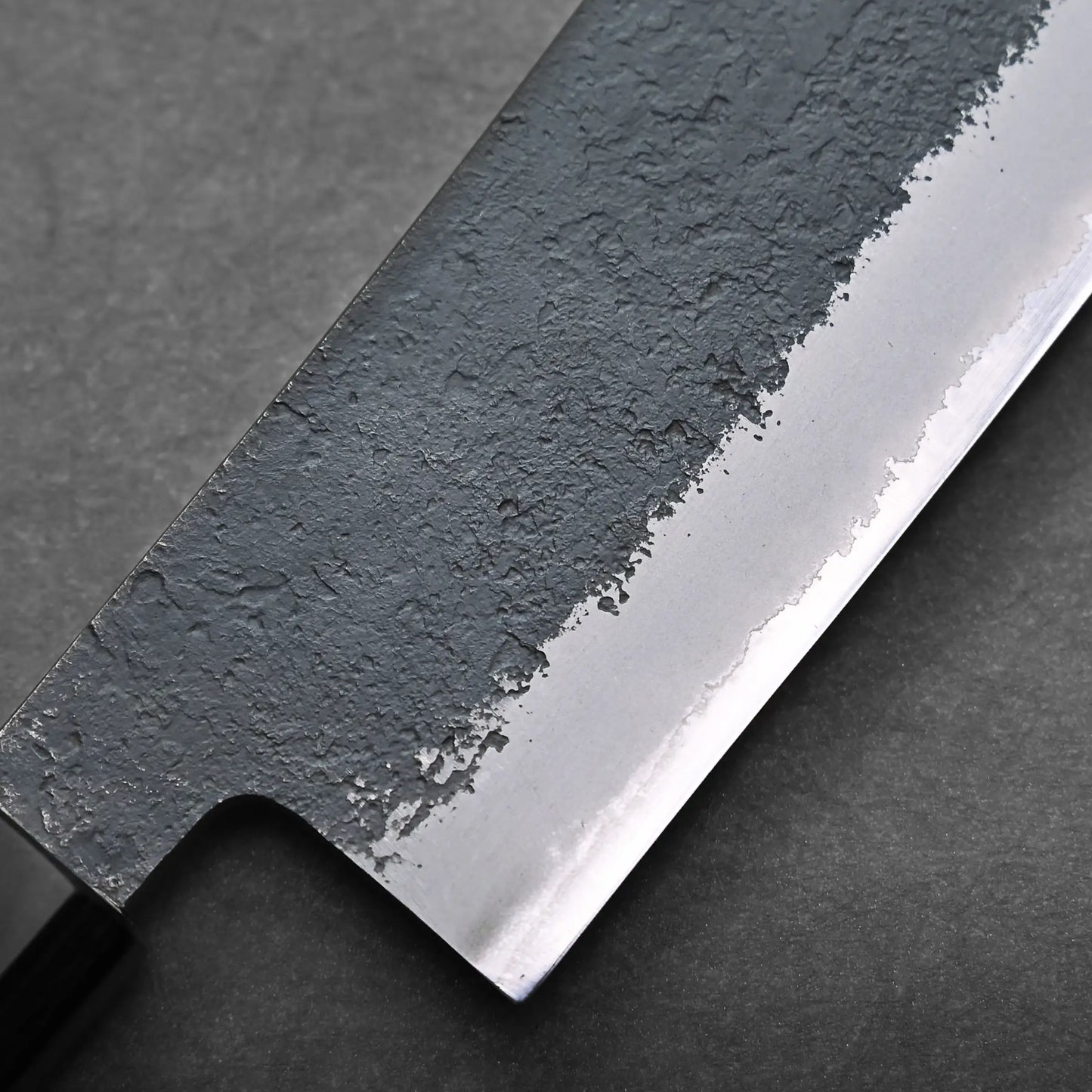 Close-up of a hakata knife blade displaying a textured, hammered finish on the upper part and a smooth, polished edge. The background is dark and out of focus.