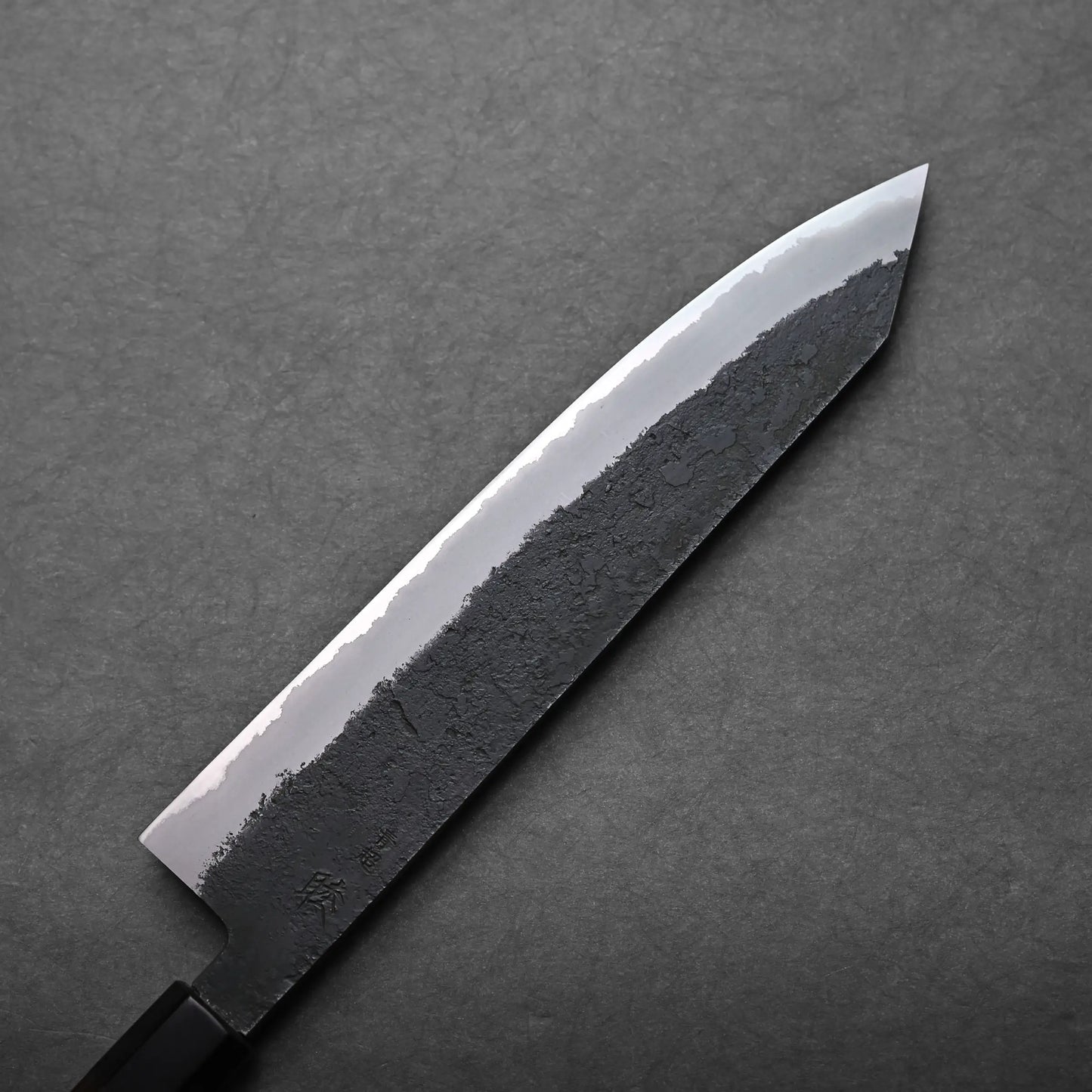 A kiritsuke Japanese chef's knife with a black textured blade and polished edge is shown on a dark gray background. The knife features a straight handle and an angled tip.