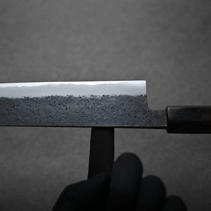 A close-up of a person wearing a black glove holding a kiritsuke Japanese chef's knife with a textured, dark blade and polished edge against a gray background. It shows the balance point.