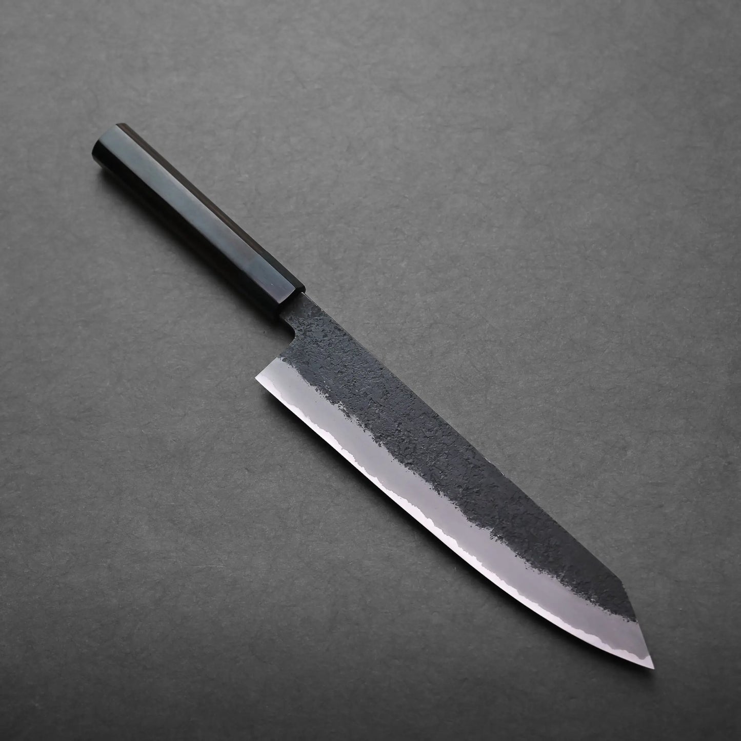 A kiritsuke gyuto with a rectangular black handle and a broad, partially textured blade rests on a dark, flat surface.