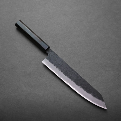 A kiritsuke gyuto with a rectangular black handle and a broad, partially textured blade rests on a dark, flat surface.