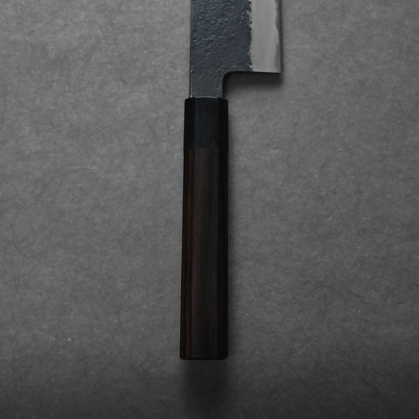 A close-up view of the handle and part of the blade of a kiritsuke gyuto placed on a dark, textured surface. The wooden handle and matte blade reflect the elegance of this Japanese chef's knife.