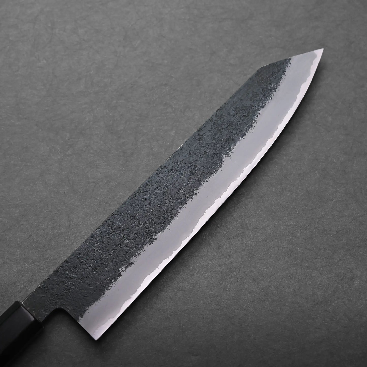 A kiritsuke gyuto featuring a textured, blackened upper blade and a sharp, polished edge rests on a dark gray surface.