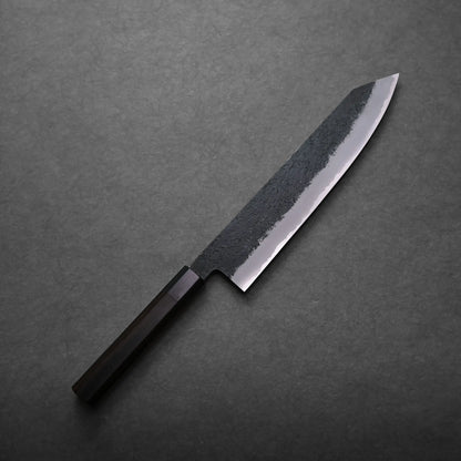 A Mutsumi Hinoura kurouchi nashiji AS kiritsuke gyuto 240mm with a dark wooden handle and a partially polished blade featuring a textured, blackened upper section, rests on a dark gray surface.