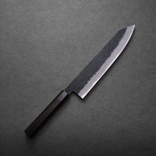 A Mutsumi Hinoura kurouchi nashiji AS kiritsuke gyuto 240mm with a dark wooden handle and a partially polished blade featuring a textured, blackened upper section, rests on a dark gray surface.